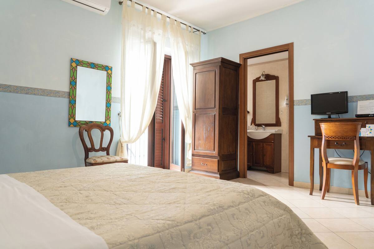 A bedroom with a bed, a wooden wardrobe, a mirror, a desk with a chair, and a television.
