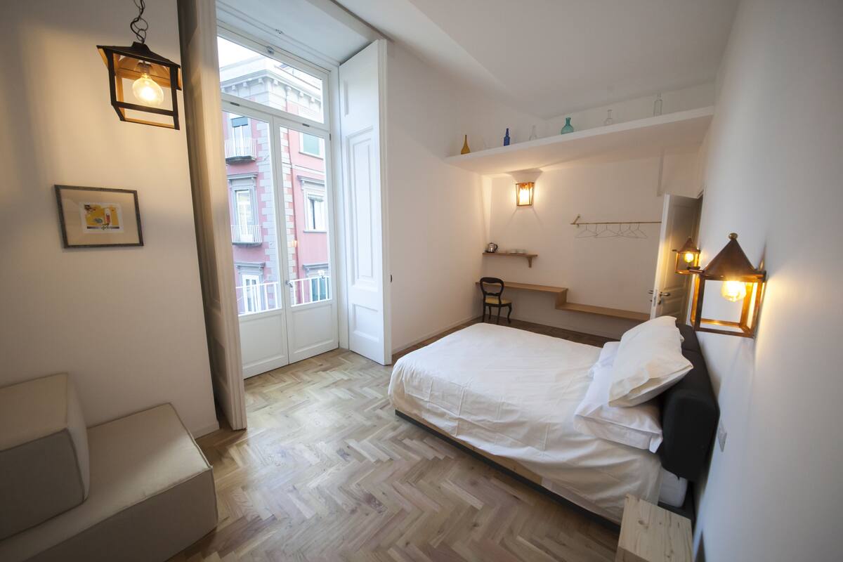 A bedroom with a bed, a chair, a bedside table, a shelf with bottles, and a window with a view of buildings.