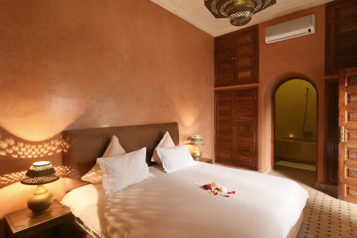 A hotel room with a large bed, a nightstand with a lamp, wooden cabinets, and an air conditioning unit.
