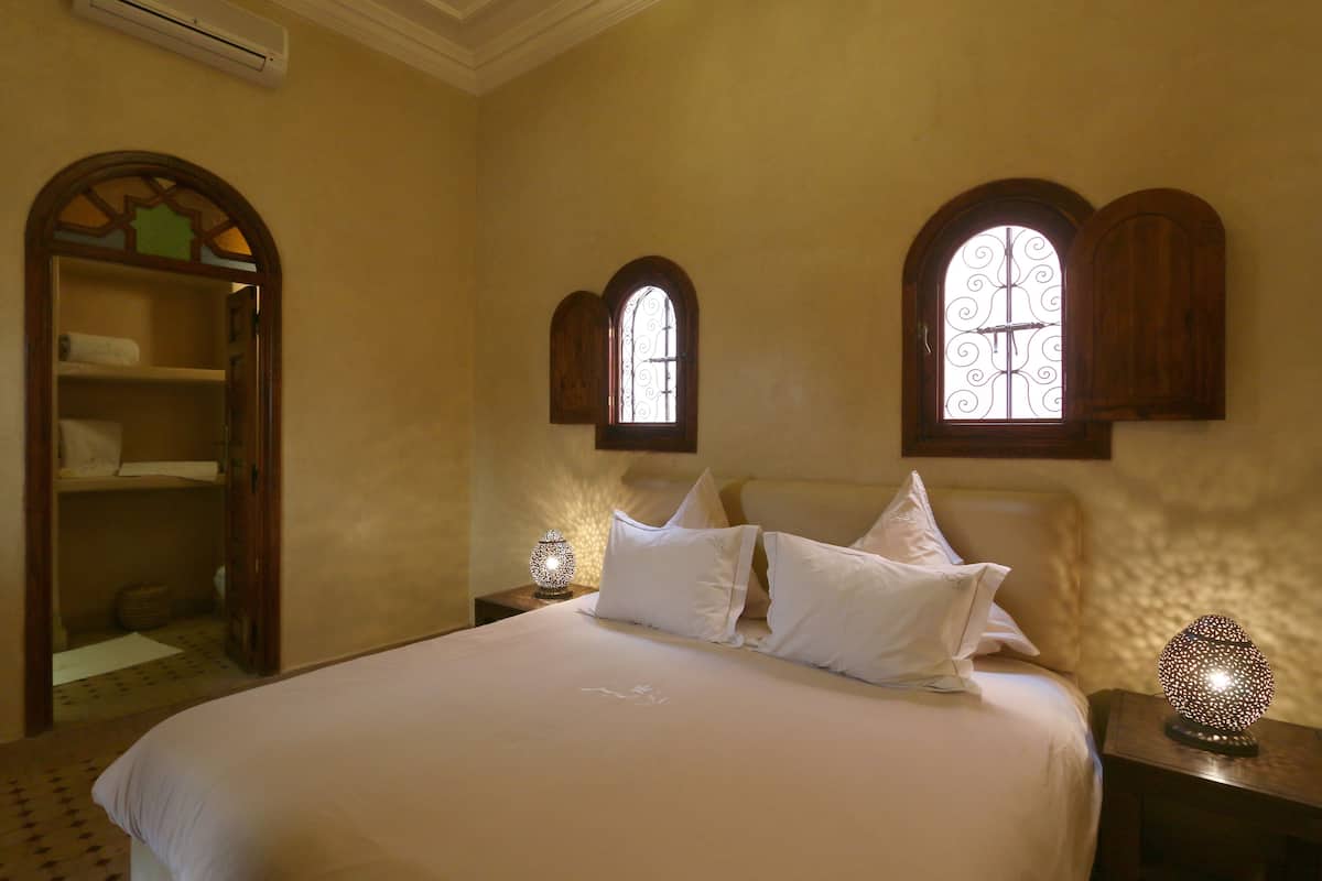 A neatly made bed with white linens, two nightstands with decorative lamps, and a wooden arched window with a metal design.