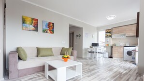 Un salon moderne avec un canapé, une table basse et des œuvres d’art accrochées au mur. On aperçoit une cuisine avec un lave-linge à l’arrière-plan.