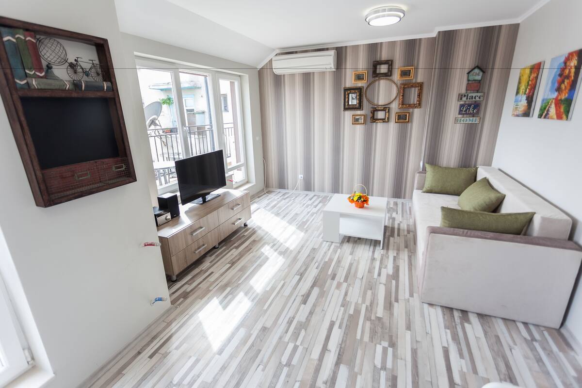 A modern living room with a white sofa, a coffee table, and a TV. There is a window with a view of the outside.