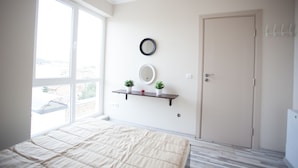 Une chambre avec une grande fenêtre, un lit, une porte, une étagère fixée au mur avec des plantes et un miroir.