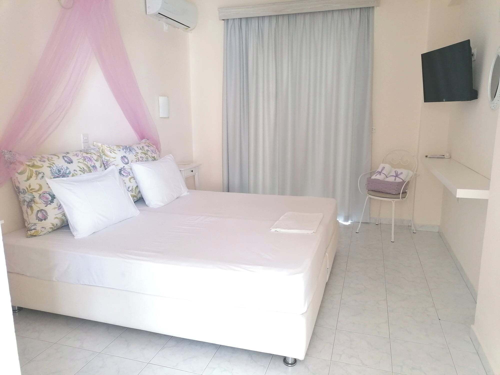 A neatly arranged bedroom with a bed, a bedside table, a chair, a TV mounted on the wall, and a window with curtains.