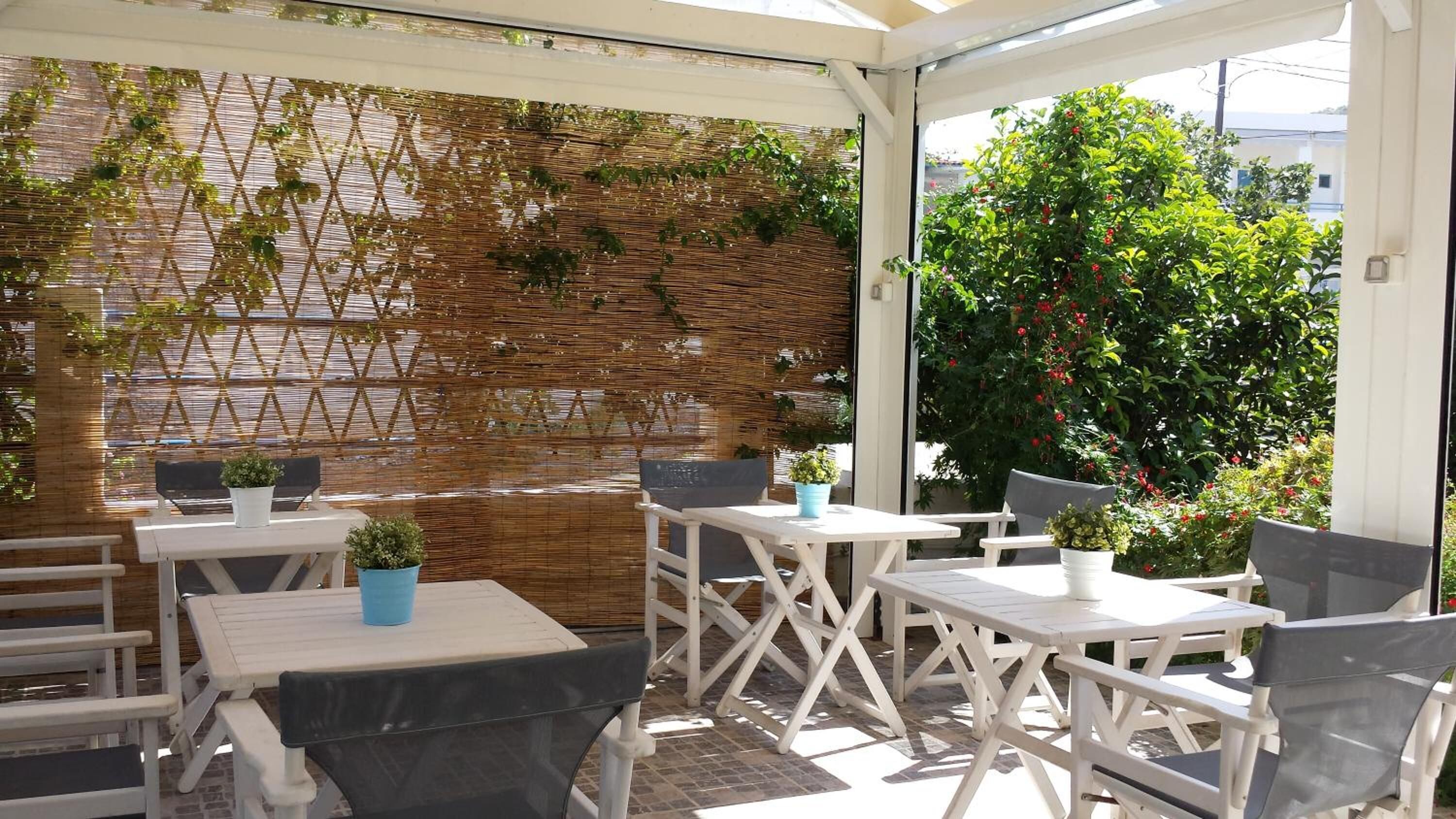 Um pátio externo com mesas e cadeiras brancas, uma parede de bambu trançado e plantas em vasos.