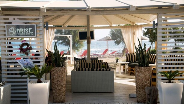 A covered outdoor seating area with wicker furniture, potted plants, and a sign for Sunrise Cocktail Bar & Cafe. - Akrogiali Hotel (Agistri)