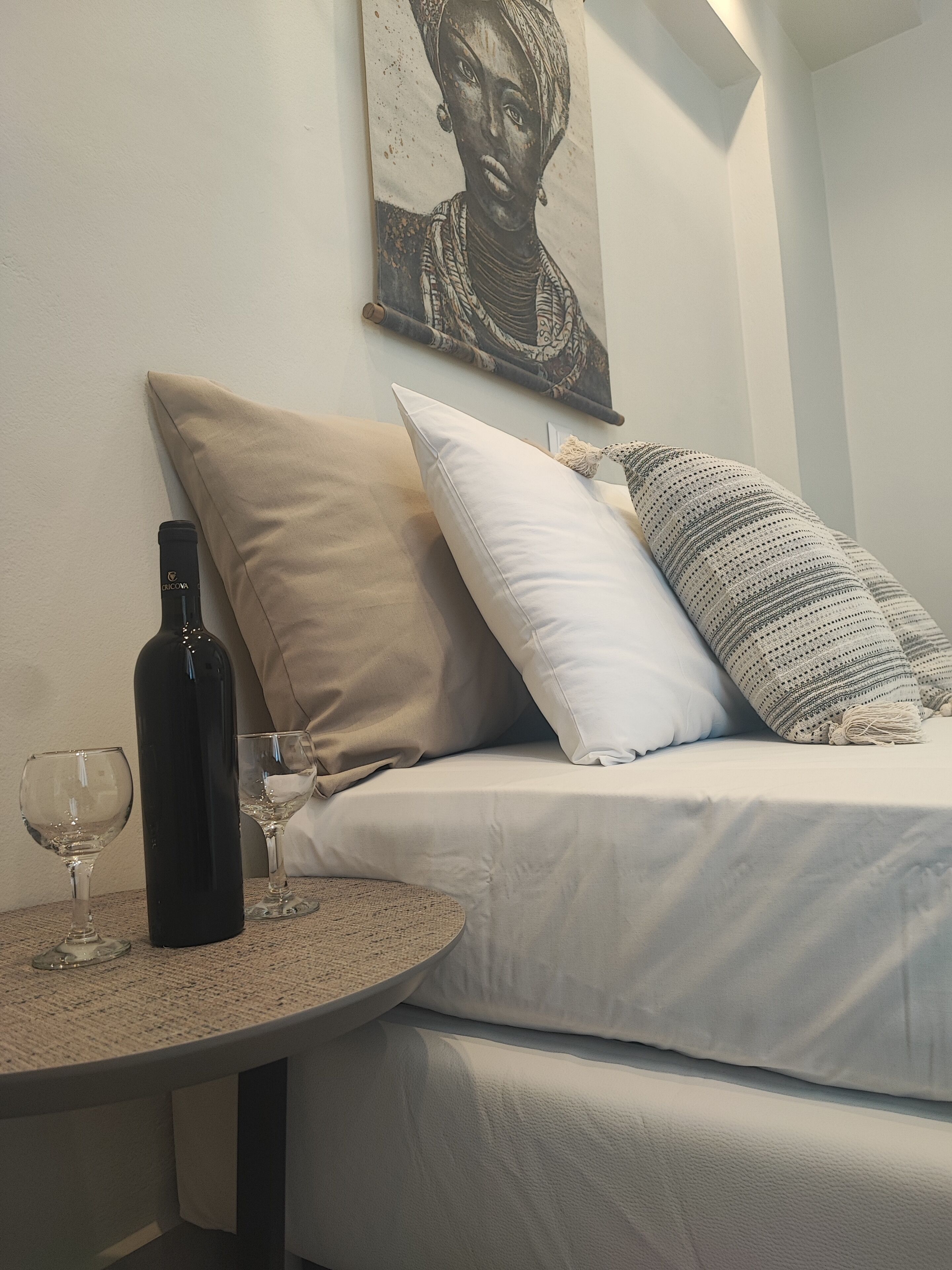 A neatly arranged bedroom with a bed, pillows, a wine bottle, and glasses on a round table.