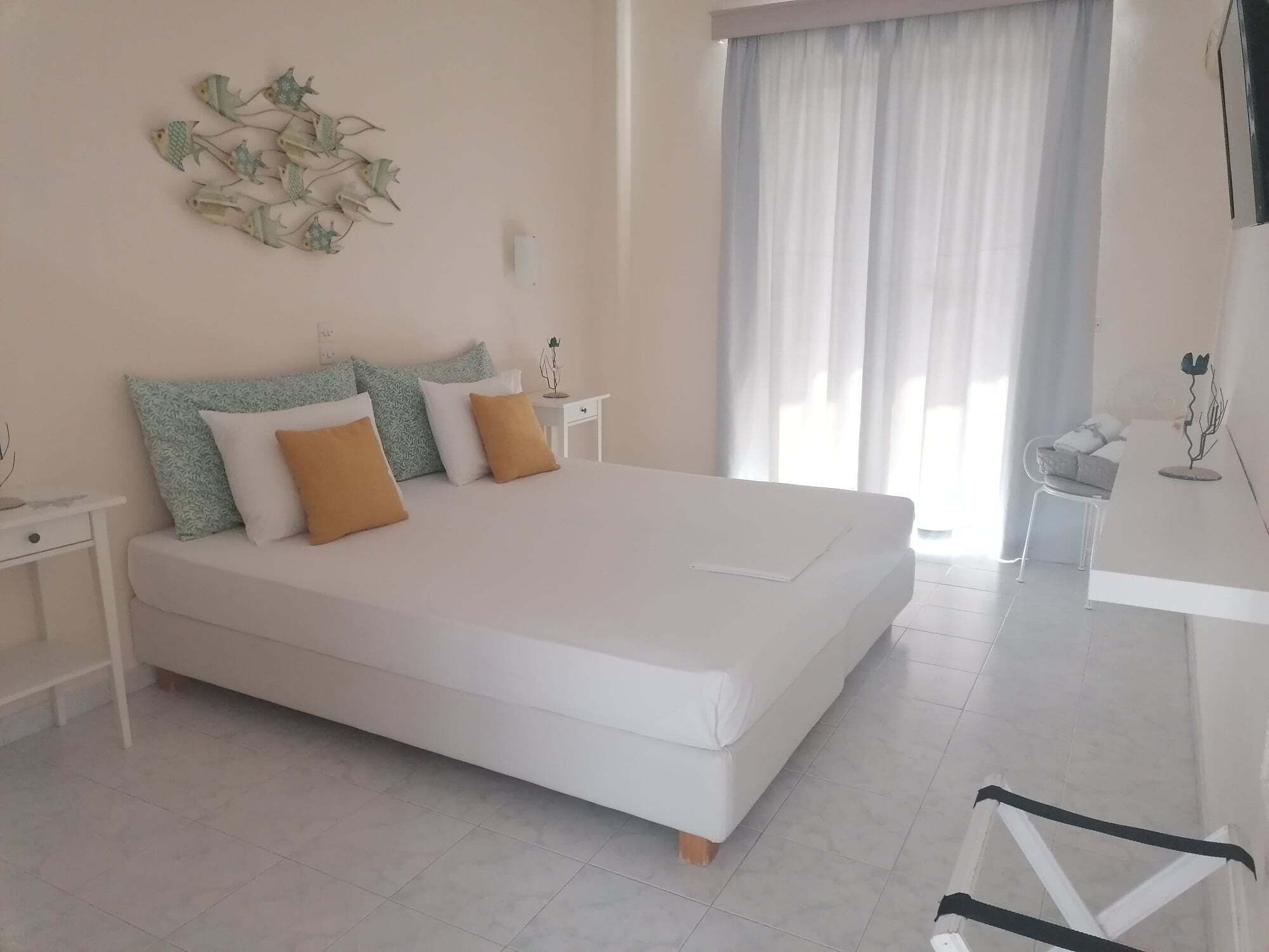 A bedroom with a large bed, white walls, a window with curtains, a bedside table, and a small plant.