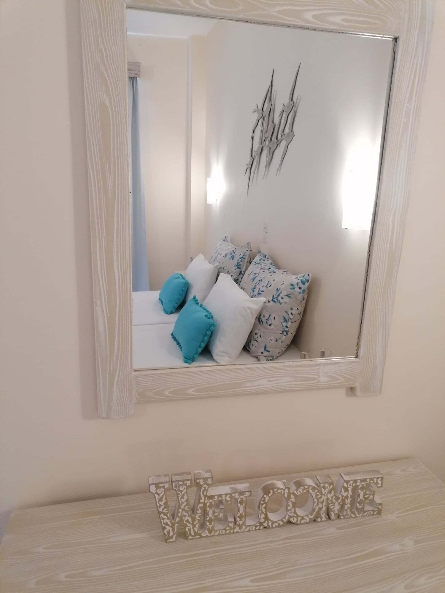 Un dormitorio con cama, almohadas, un espejo decorativo y un cartel que dice 'WELCOME'.