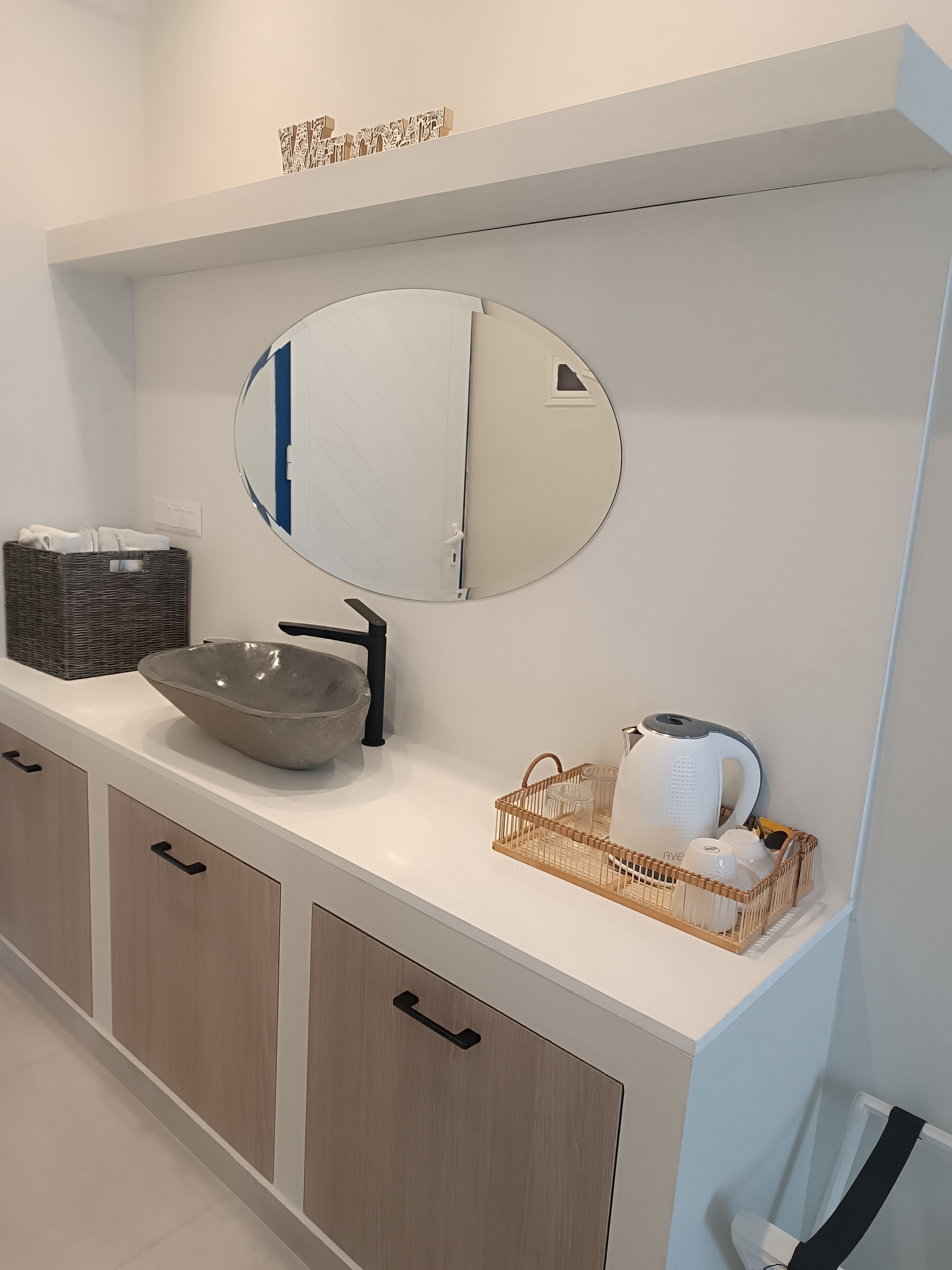 A modern bathroom with a large mirror, a bowl sink, a towel basket, and a tea kettle on the counter.
