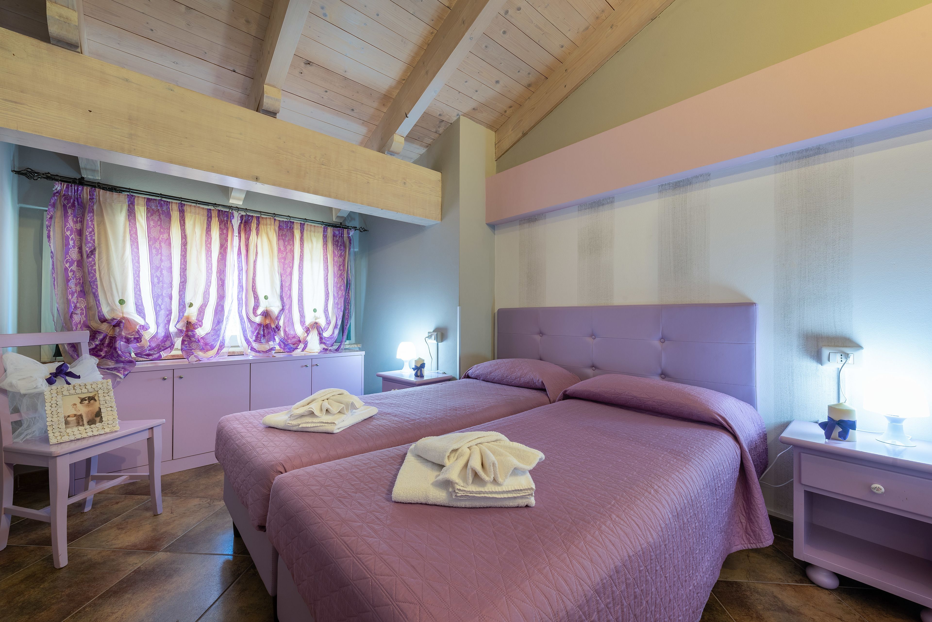 A bedroom with two beds, wooden ceiling beams, a window with purple curtains, and a bedside table with a lamp.