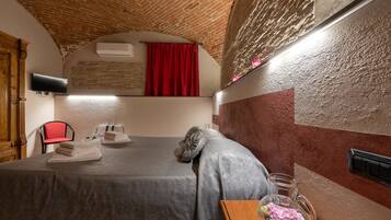 Habitación de hotel con techo de ladrillo, cama con cubrecama gris, una silla roja, una mesita de noche de madera con una jarra de vidrio y un aire acondicionado de pared.