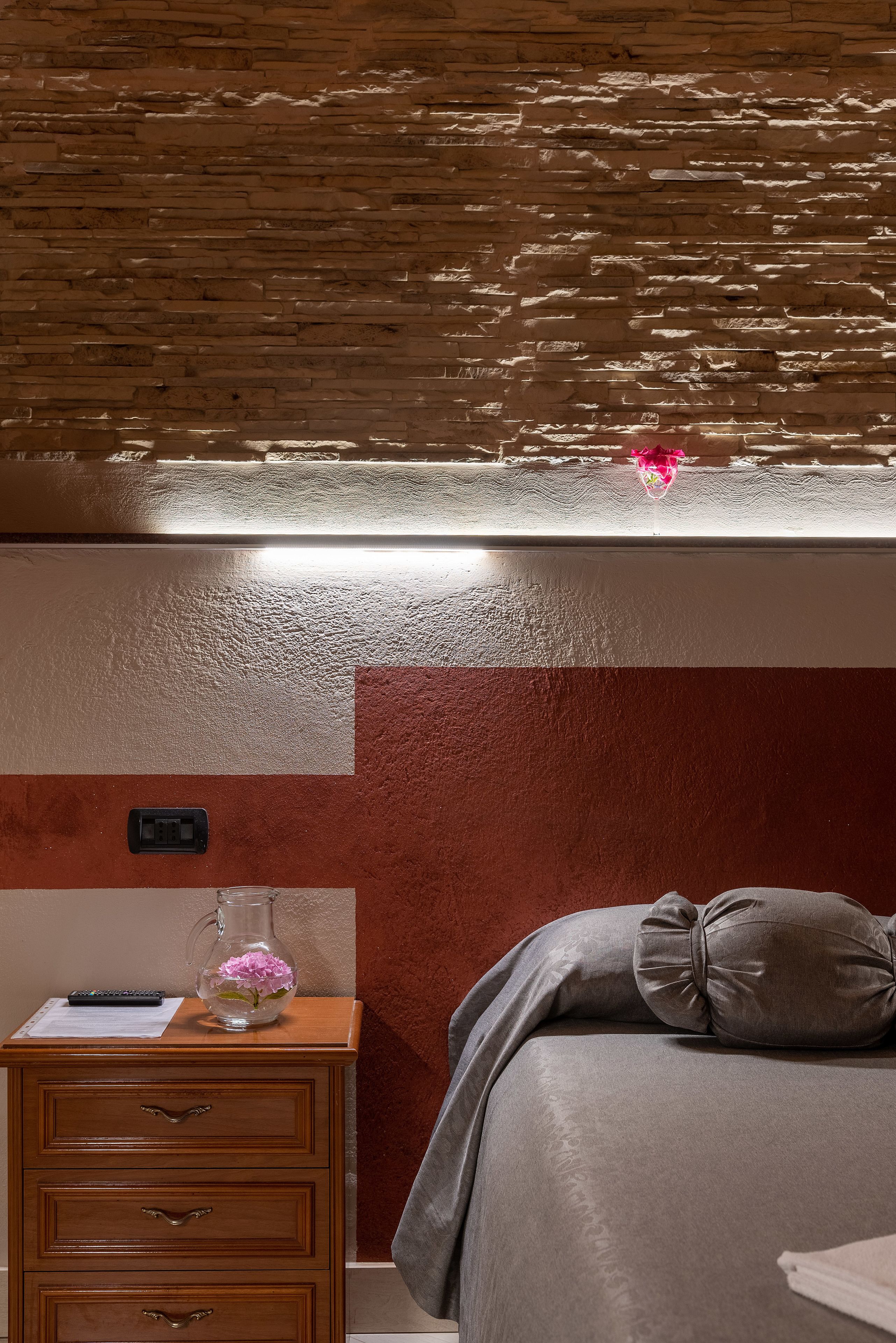 Une chambre d’hôtel avec un mur à texture bois, une table de chevet avec une télécommande, un vase avec des fleurs et un lit recouvert d’une couverture grise et d’un oreiller.