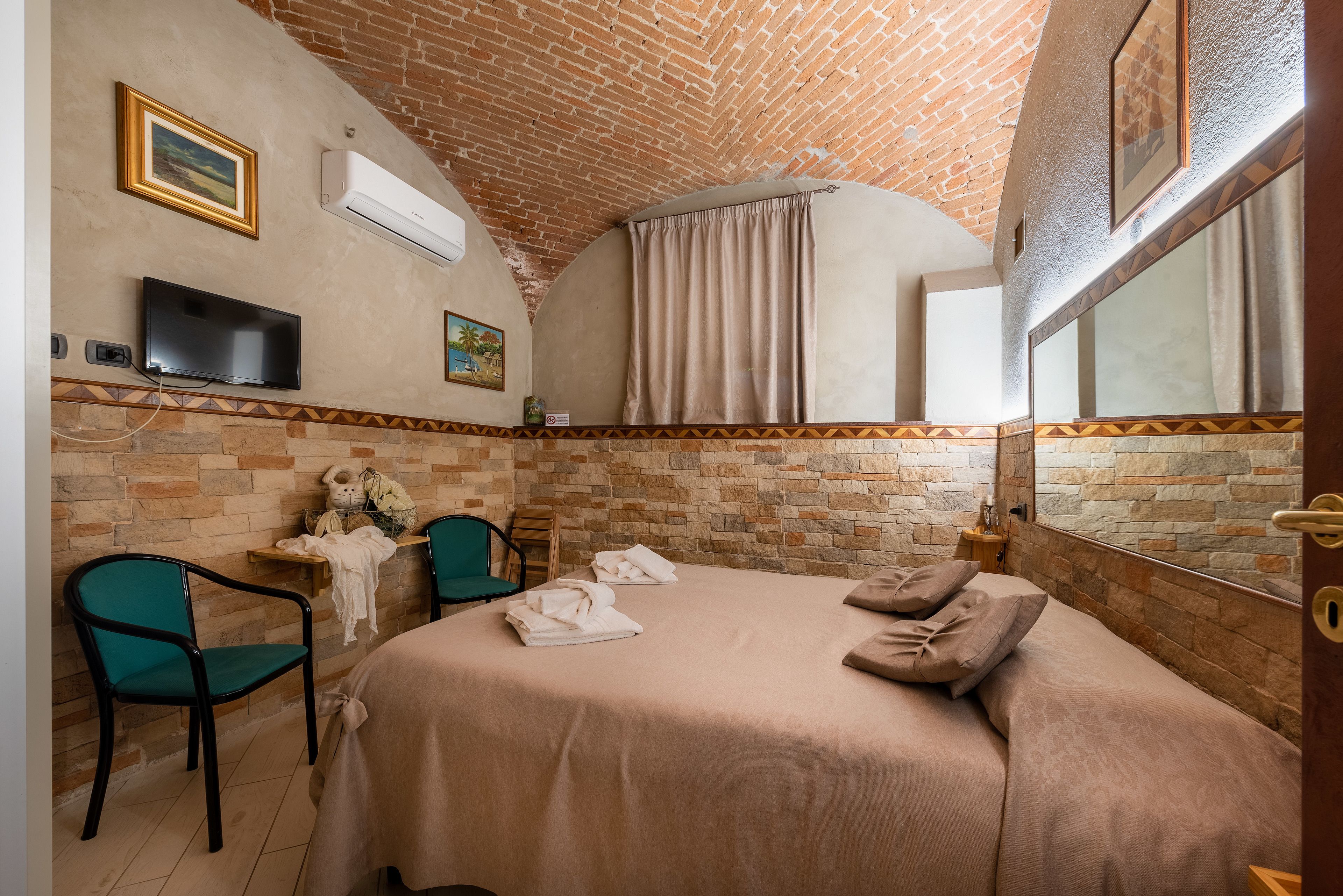 A hotel room with a brick ceiling, a flat-screen TV, a bed with towels and pillows, a chair, and a small table with a lamp.