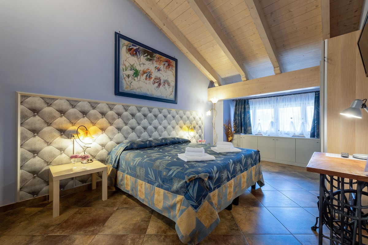 Une chambre à coucher avec une tête de lit capitonnée, un lit recouvert d’une couvre-lit à motifs floraux, une table de chevet avec une lampe et une fenêtre avec des rideaux.