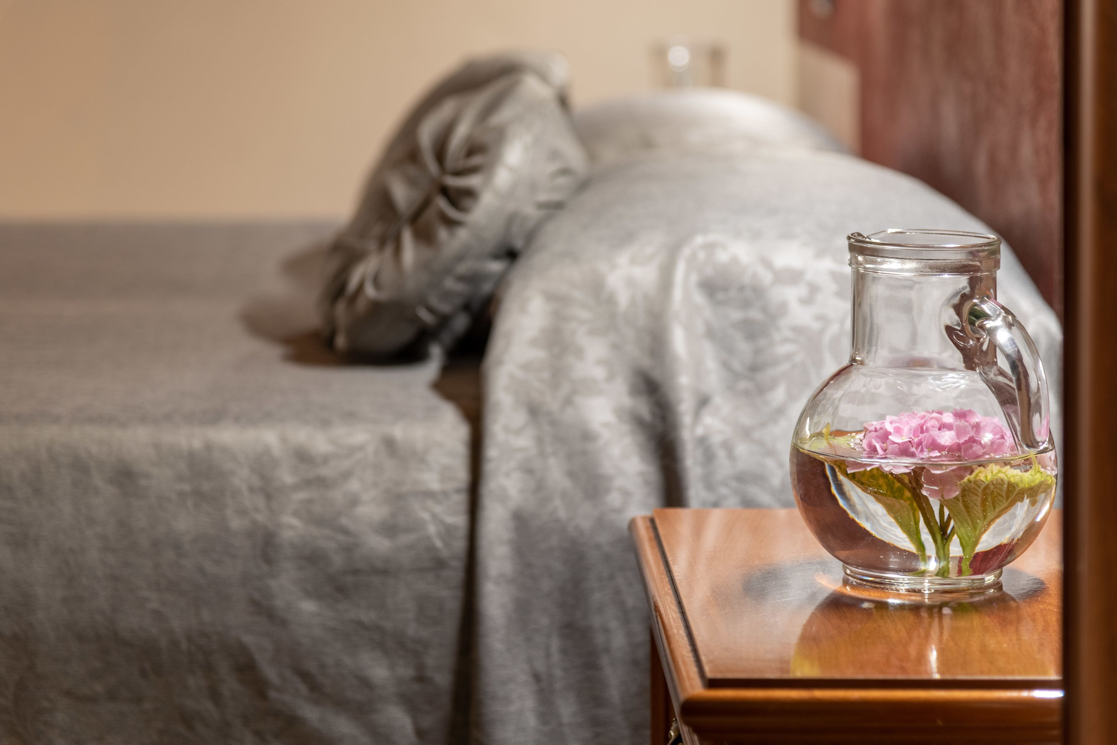 A bedroom with a bed, a wooden nightstand, and a clear glass vase with pink flowers.