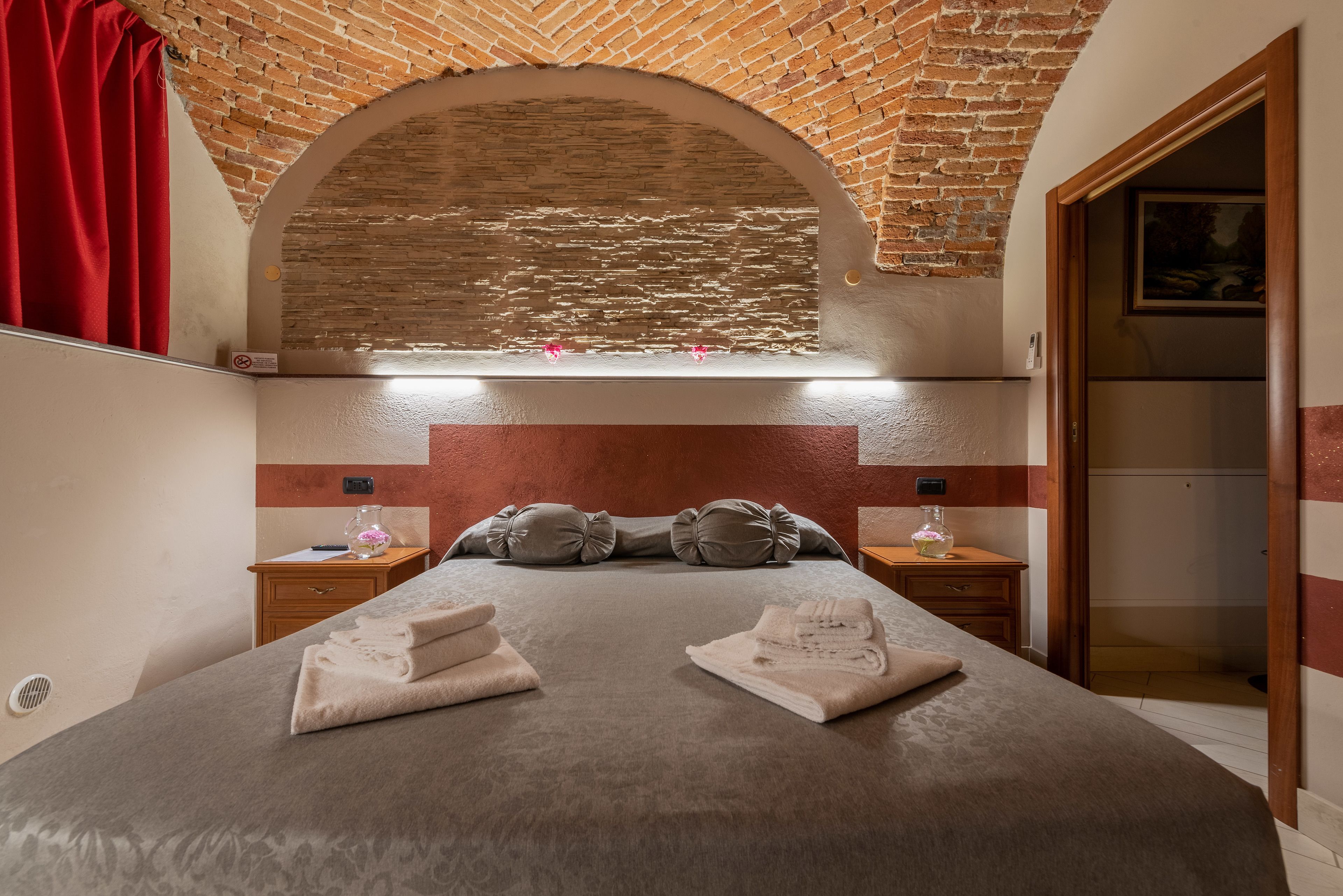 Une chambre d’hôtel avec un plafond en briques, un lit avec des serviettes soigneusement pliées, une table de chevet avec un vase et un rideau rouge.