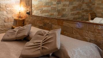 Una cama con ropa de cama y almohadas color beige, una mesita de noche con una lámpara, y una pared de piedra con un diseño geométrico.
