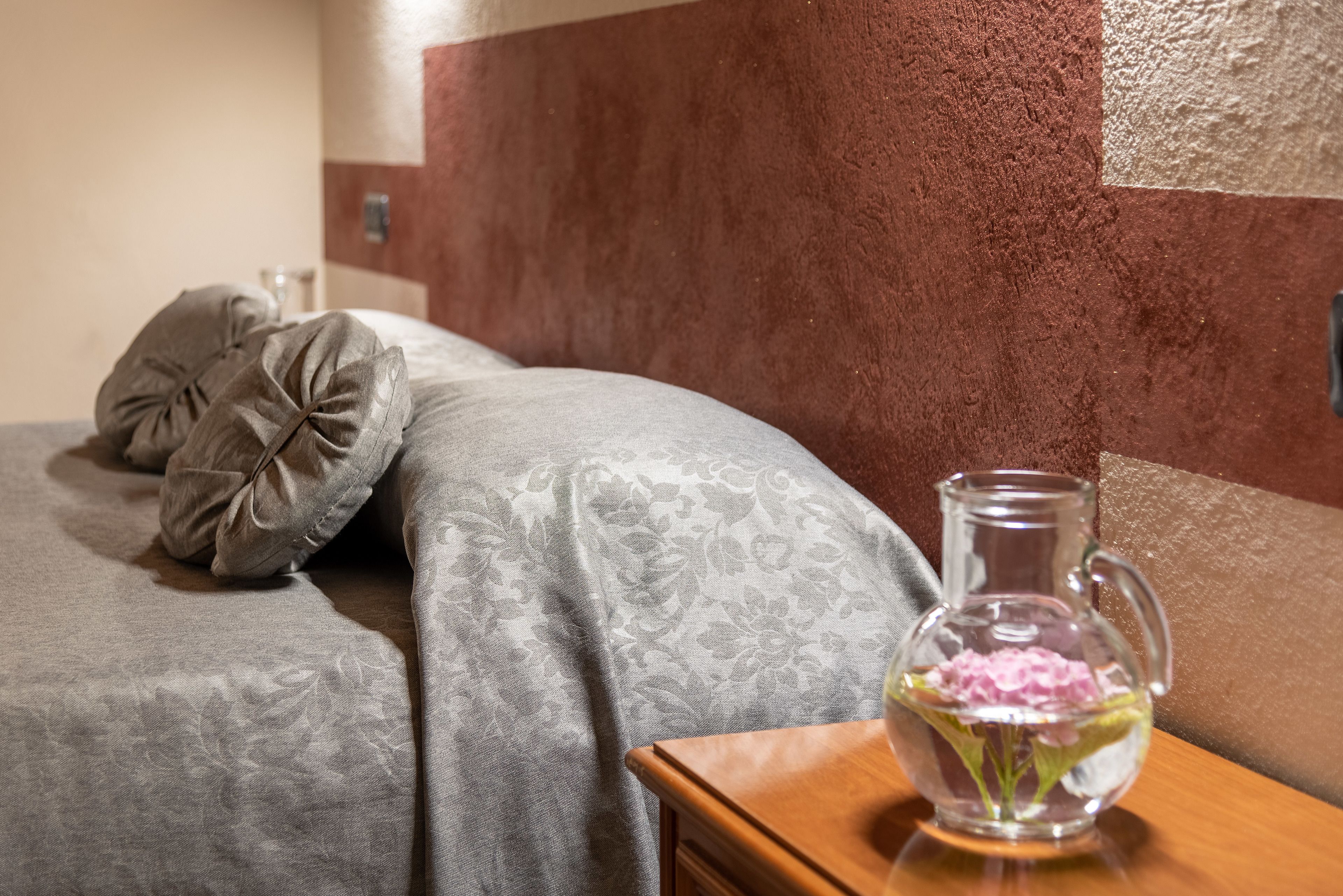 Una cama con ropa de cama gris y dos almohadas, un florero de vidrio transparente con flores rosadas sobre una mesita de noche de madera.