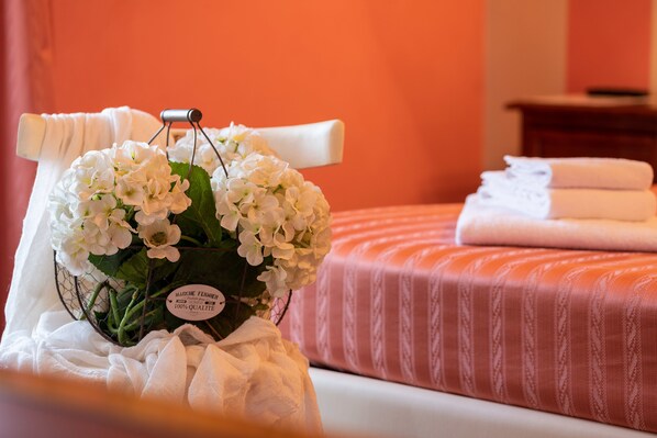 Una cama bien tendida con una colcha a rayas rosa y blanca, una toalla blanca sobre la cama y un ramo de flores blancas en una cesta de alambre.