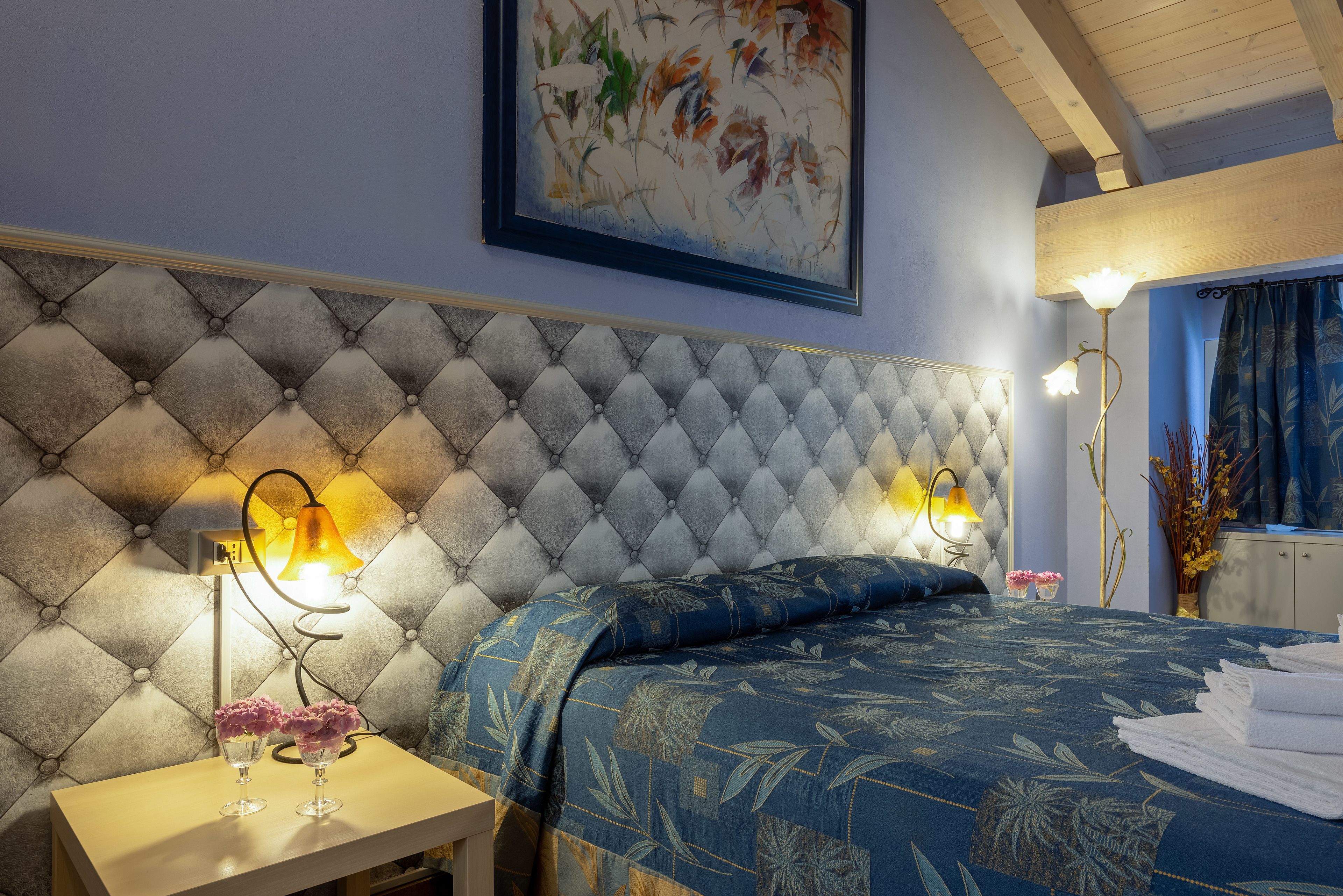 A bedroom with a tufted headboard, two bedside lamps, a bed with a floral bedspread, and a painting on the wall.