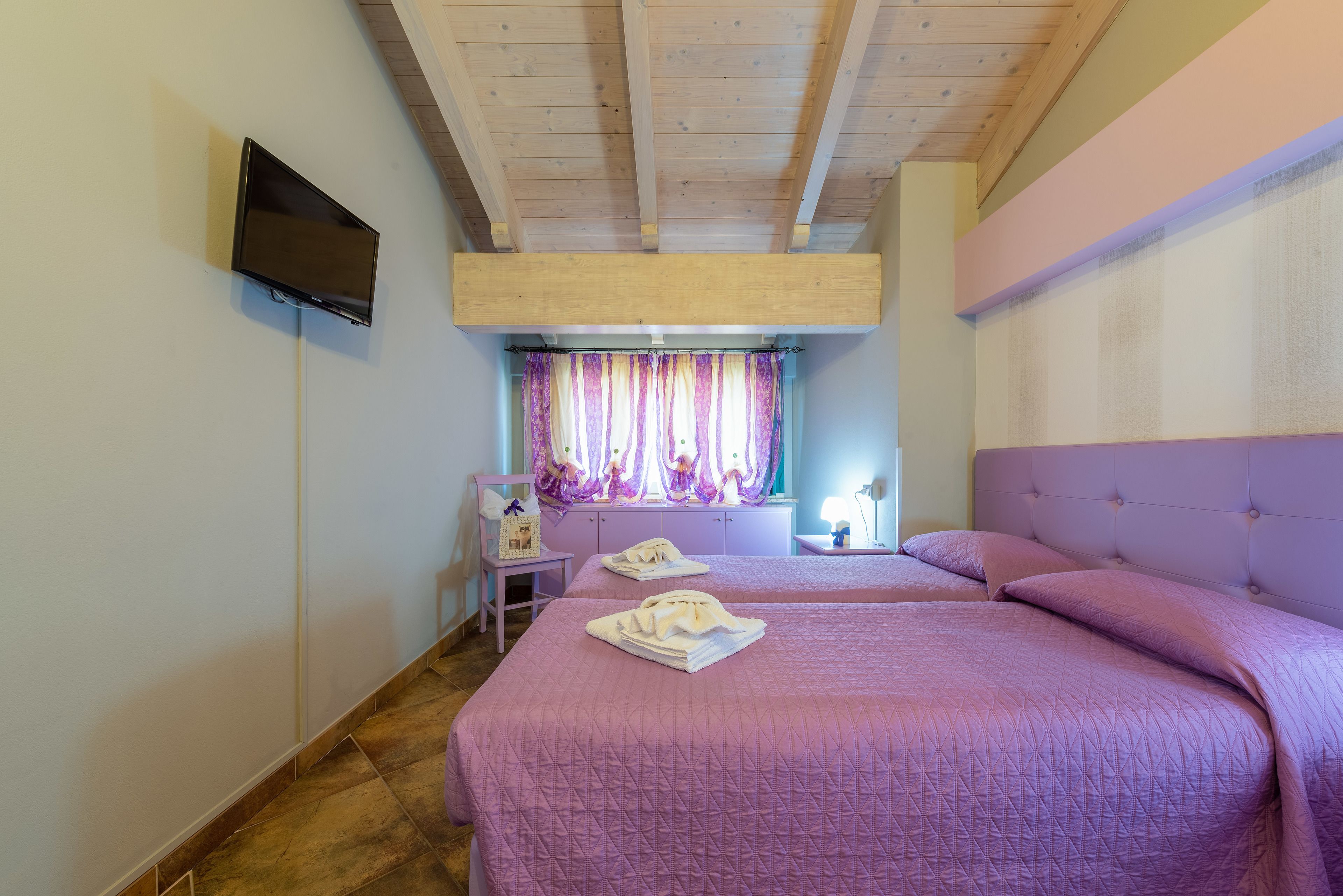 Un dormitorio con dos camas, un televisor montado en la pared, un ventanal con cortinas y techo de madera.