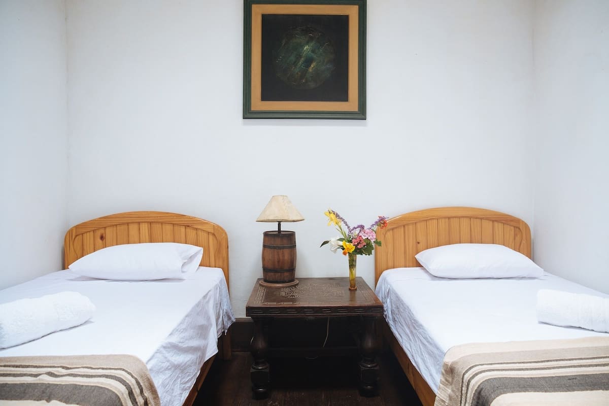 Una habitación con dos camas de madera, ropa de cama blanca, una mesita con una lámpara y un jarrón con flores.