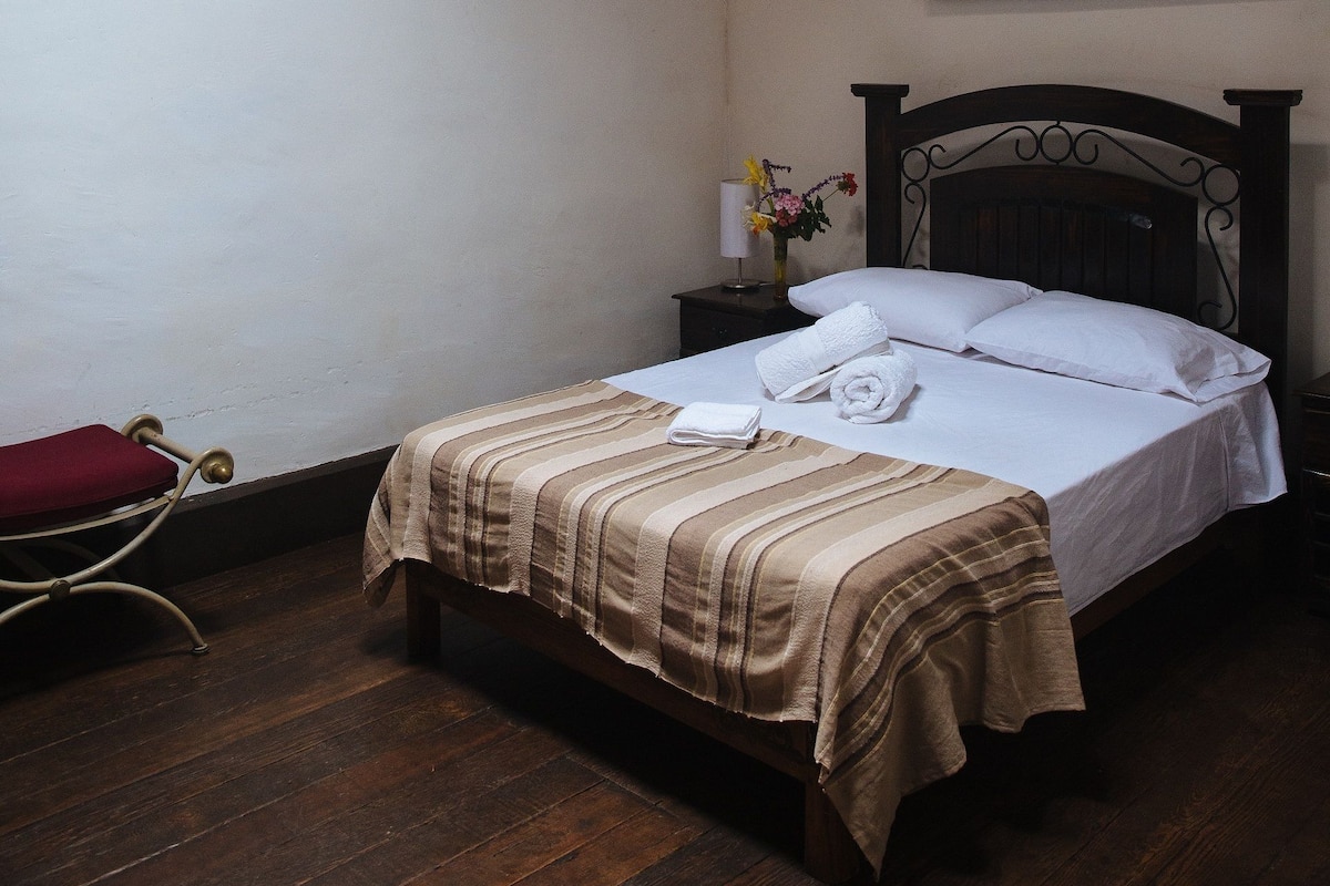 Un dormitorio con cama de madera, lámpara colgante, mesita de noche con un florero y un cuadro en la pared.