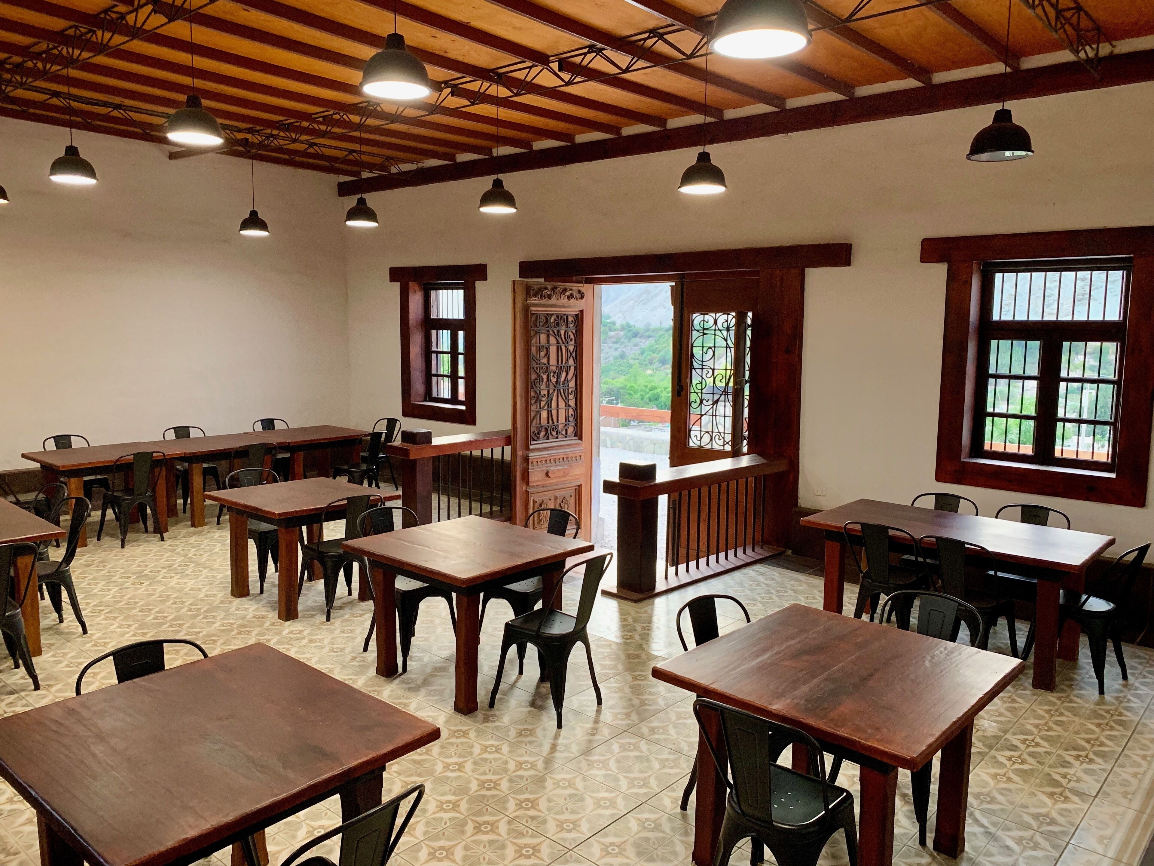 A dining area with wooden tables and chairs, large windows with decorative grills, and hanging pendant lights.