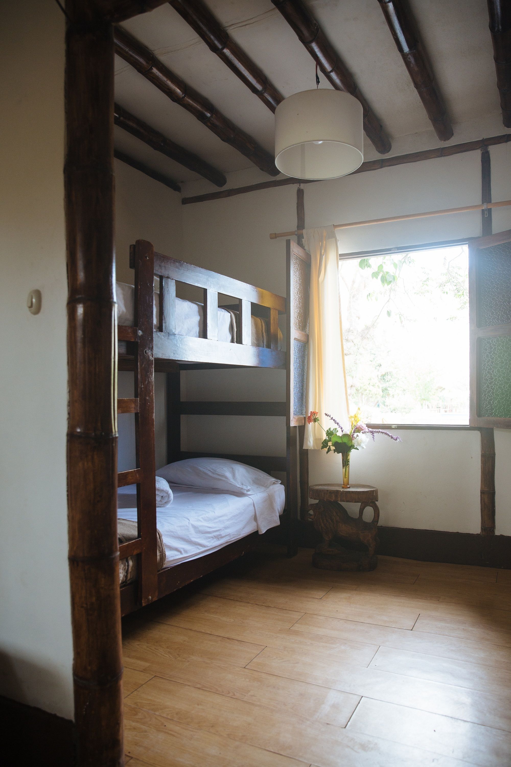 Ein Zimmer mit Etagenbett, Holzboden, einer Pendelleuchte, einem Fenster mit Vorhängen und einem kleinen Tisch mit einer Vase.