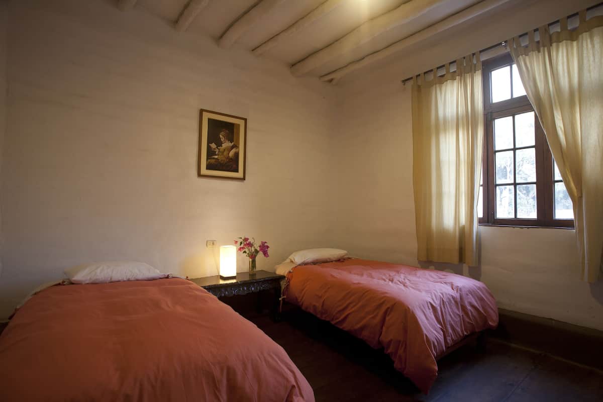 Una habitación con dos camas individuales, una mesita de noche con lámpara, un cuadro en la pared y una ventana con cortinas.
