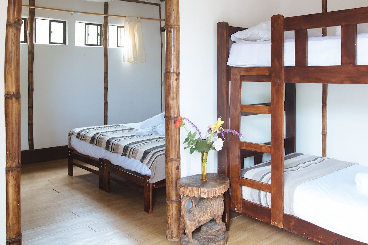 Una habitación rústica con una cama litera de madera, una mesita de noche con un florero, y piso de madera.
