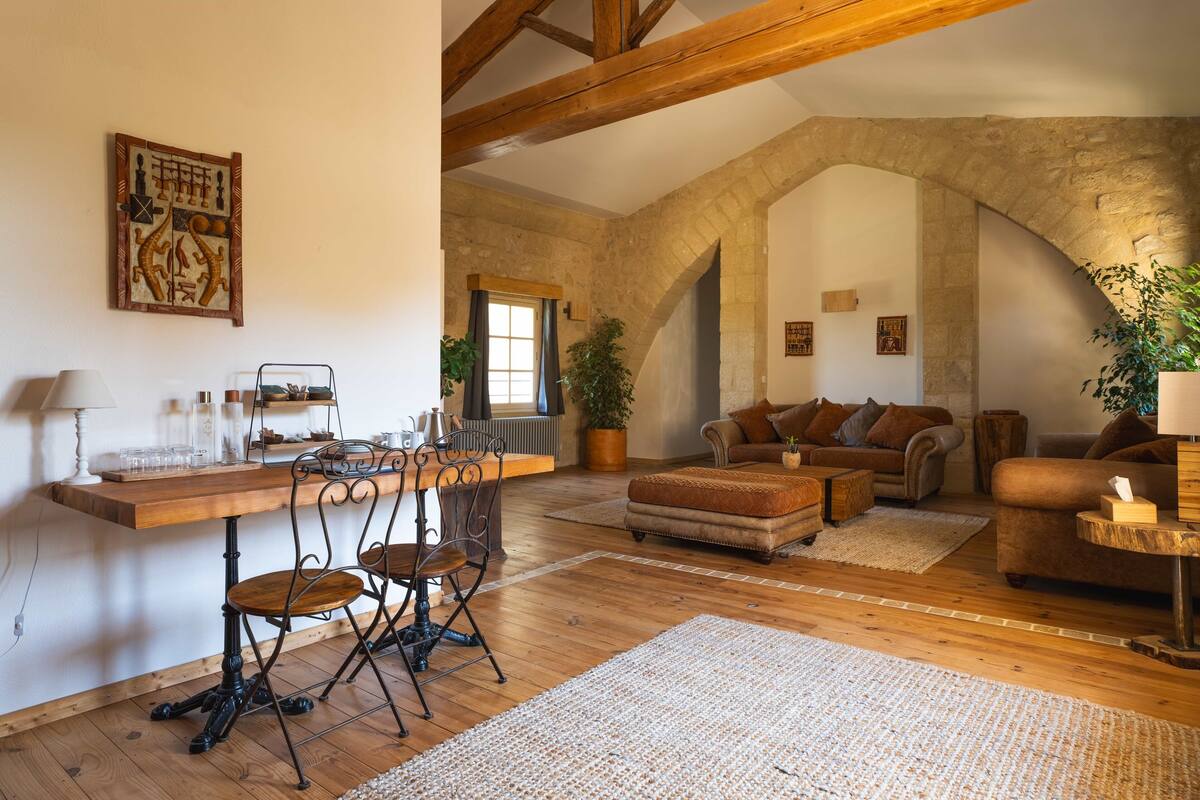 Una sala de estar rústica con una mesa de comedor de madera, sillas, un sofá y una mesa de centro. Se observa un arco de piedra y un amplio ventanal con cortinas.