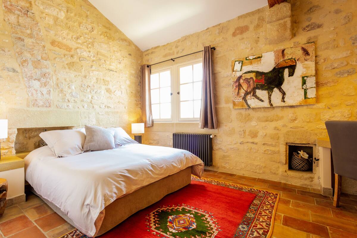 Un dormitorio con pared de piedra, cama con ropa de cama blanca, alfombra roja estampada, ventana con cortinas y chimenea con un adorno.