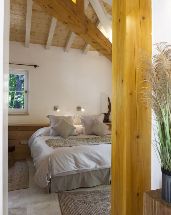 Un dormitorio con techo de madera, una cama con ropa de cama blanca, un ventanal con vistas a árboles y una planta en maceta.
