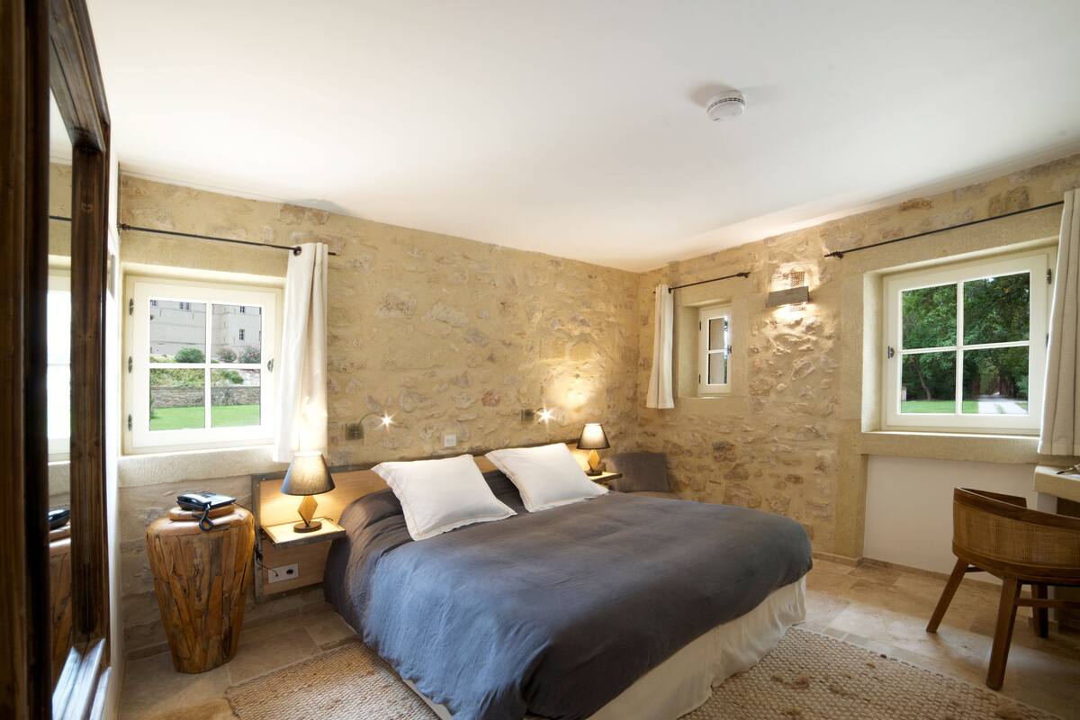 Un dormitorio con pared de piedra, una cama con manta oscura, dos lámparas de noche, una mesita de madera y una silla.