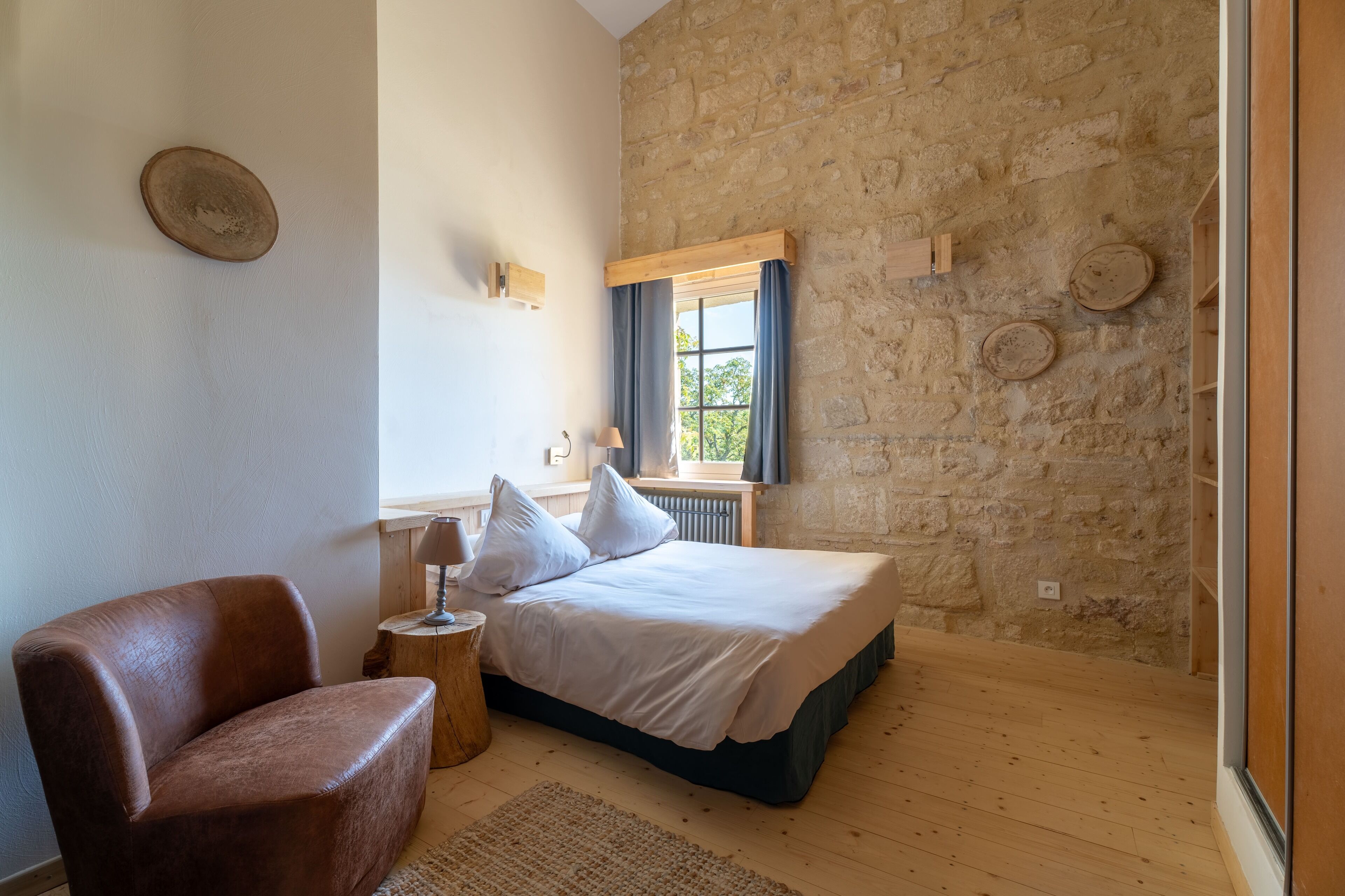 A bedroom with a stone wall, a bed with white bedding, a wooden chair, and a small wooden side table.