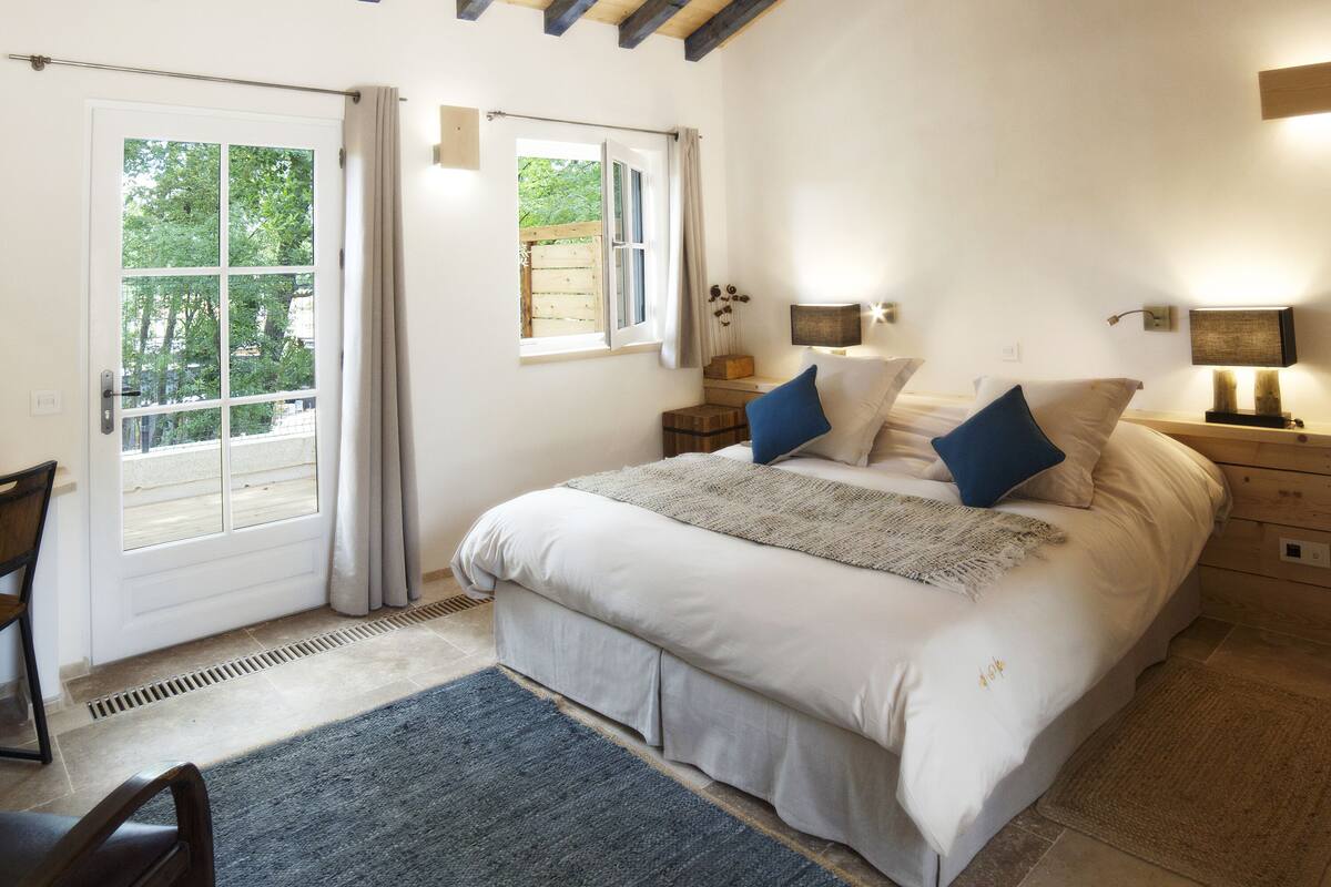 Un dormitorio con una cama grande, dos mesitas de noche con lámparas, un ventanal con cortinas y techo de madera.