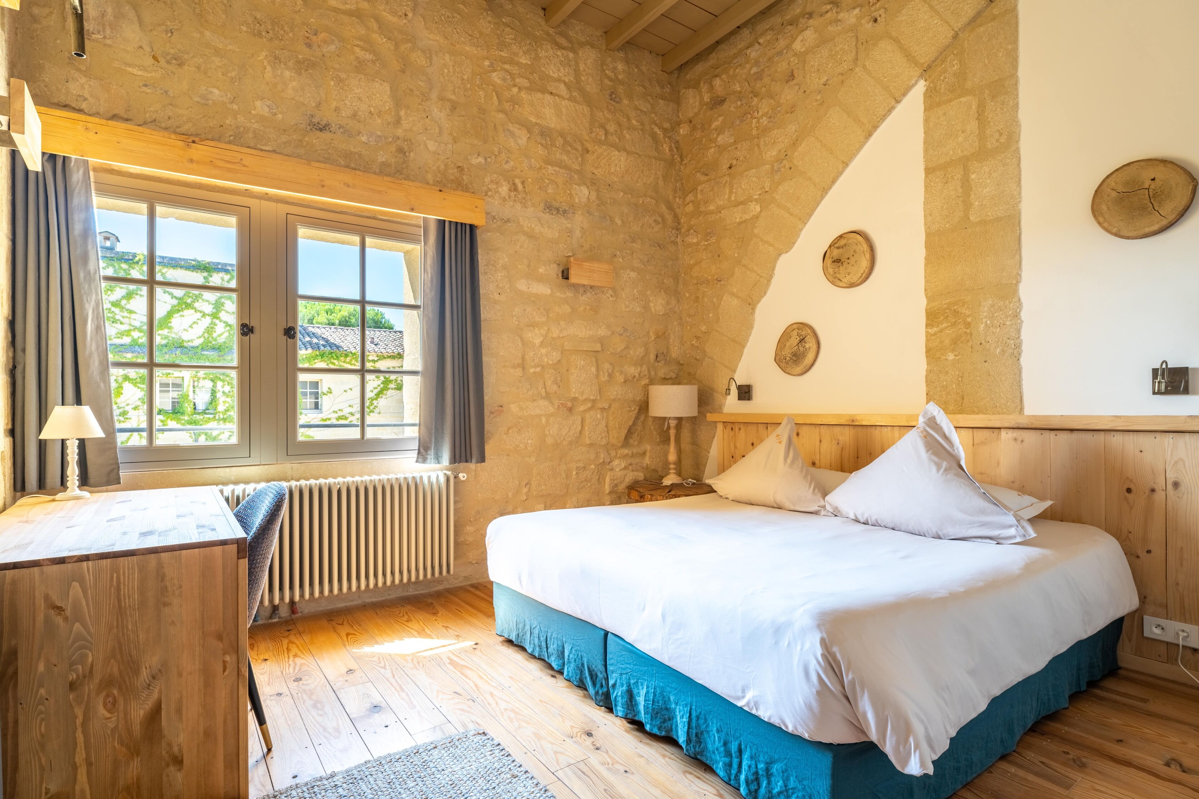 A rustic bedroom with a stone wall, a wooden bed, a desk, and a window with curtains.