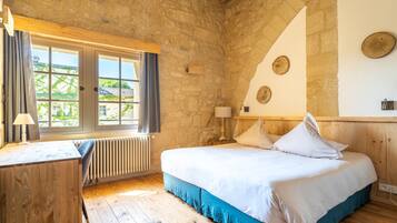 A rustic bedroom with a stone wall, a wooden bed, a desk, and a window with curtains.