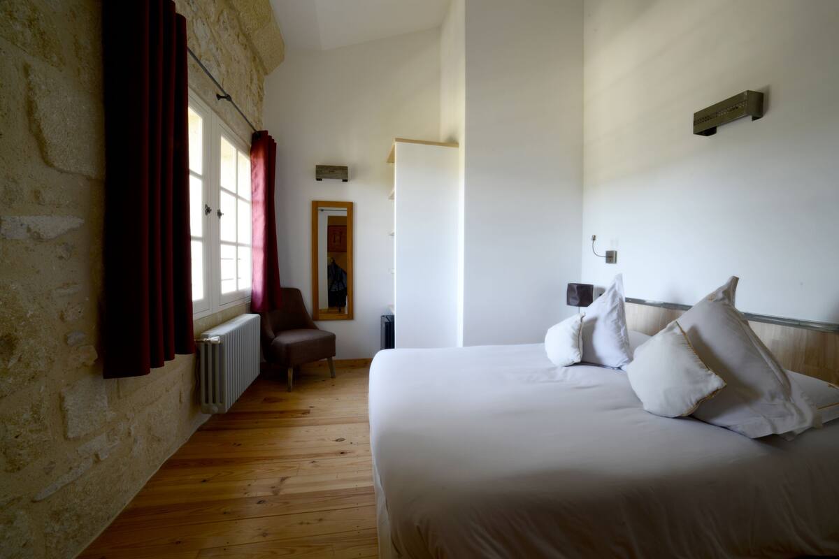 Un dormitorio con suelo de madera, una cama con ropa blanca, una silla, un radiador y una ventana con cortinas.