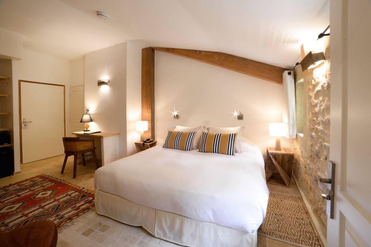 Un dormitorio con una cama grande, dos mesitas de noche con lámparas, una silla de madera, un escritorio y un tapete estampado.