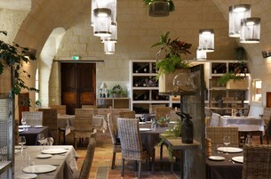 Restaurante com pé-direito alto, móveis de vime e luminárias pendentes. Mesas com toalhas brancas e copos de vinho.