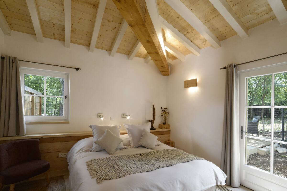 Un dormitorio con techo de madera, cama con ropa blanca, mesita de noche con lámpara y ventana con cortinas.