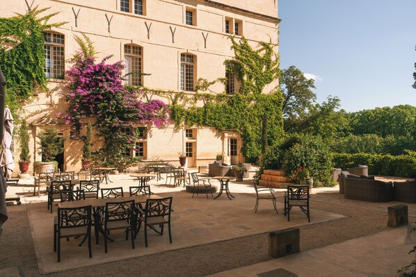 An outdoor patio with tables and chairs, surrounded by a building with large windows and greenery. - Château de Pondres (Villevieille)