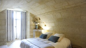 Un dormitorio con pared de piedra, una cama con ropa de cama blanca, un florero con flores y una ventana con cortinas.