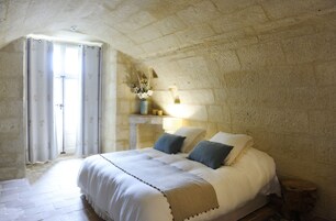 Um quarto com parede de pedra, uma cama com roupa de cama branca, um vaso com flores e uma janela com cortinas.