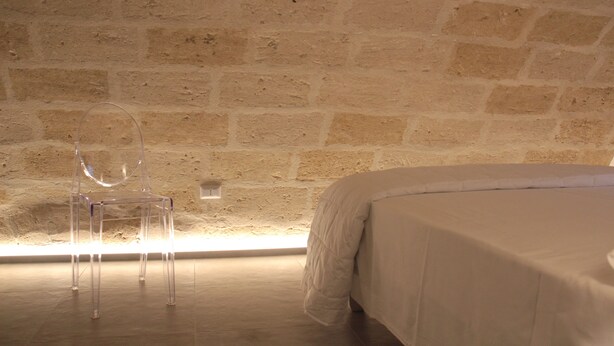 Una stanza con parete in pietra, una sedia trasparente in plastica e un letto con copriletto bianco.