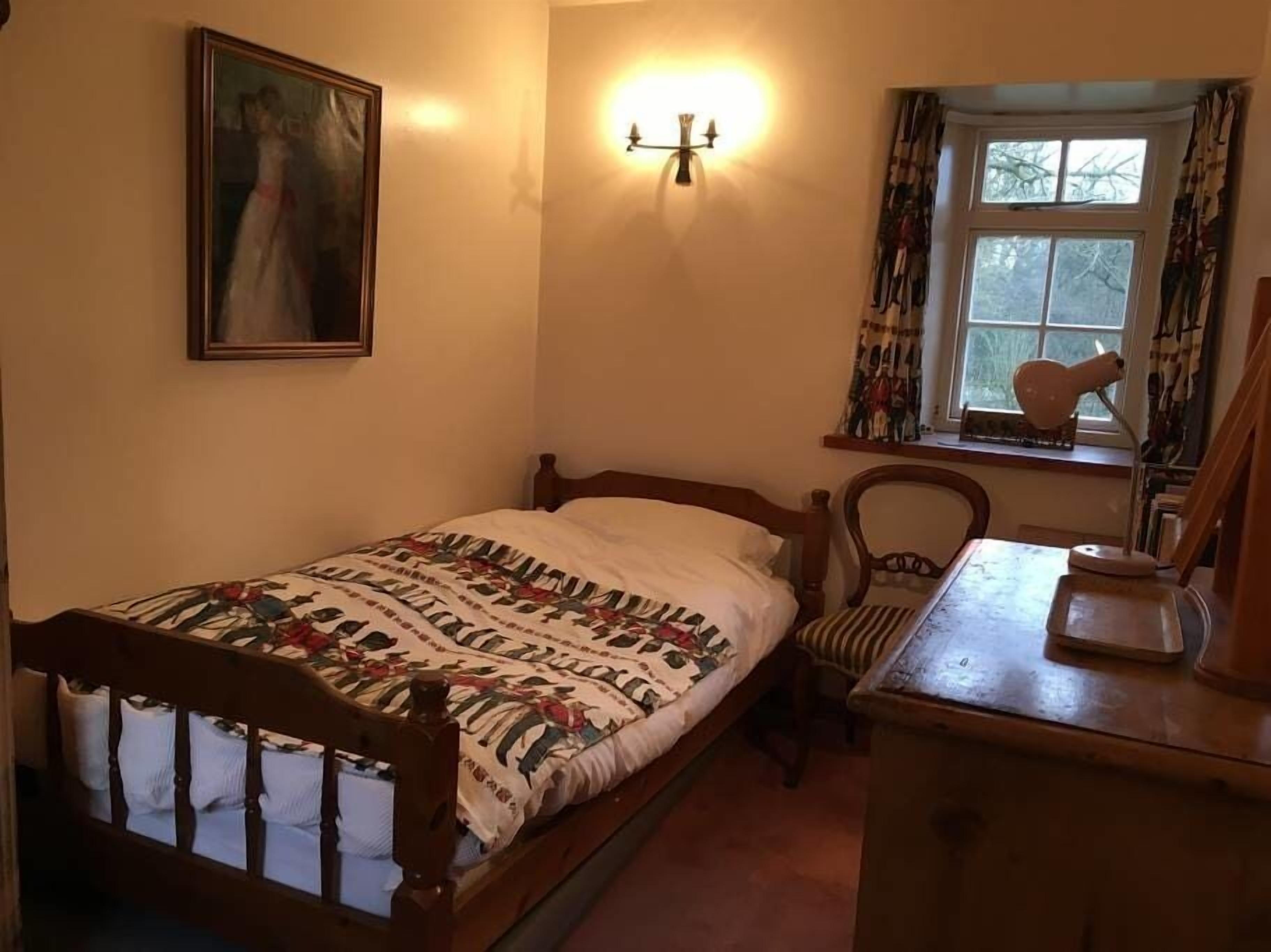Ein Schlafzimmer mit Bett, Stuhl, Kommode, Gemälde, Lampe und einem Fenster mit Vorhängen.