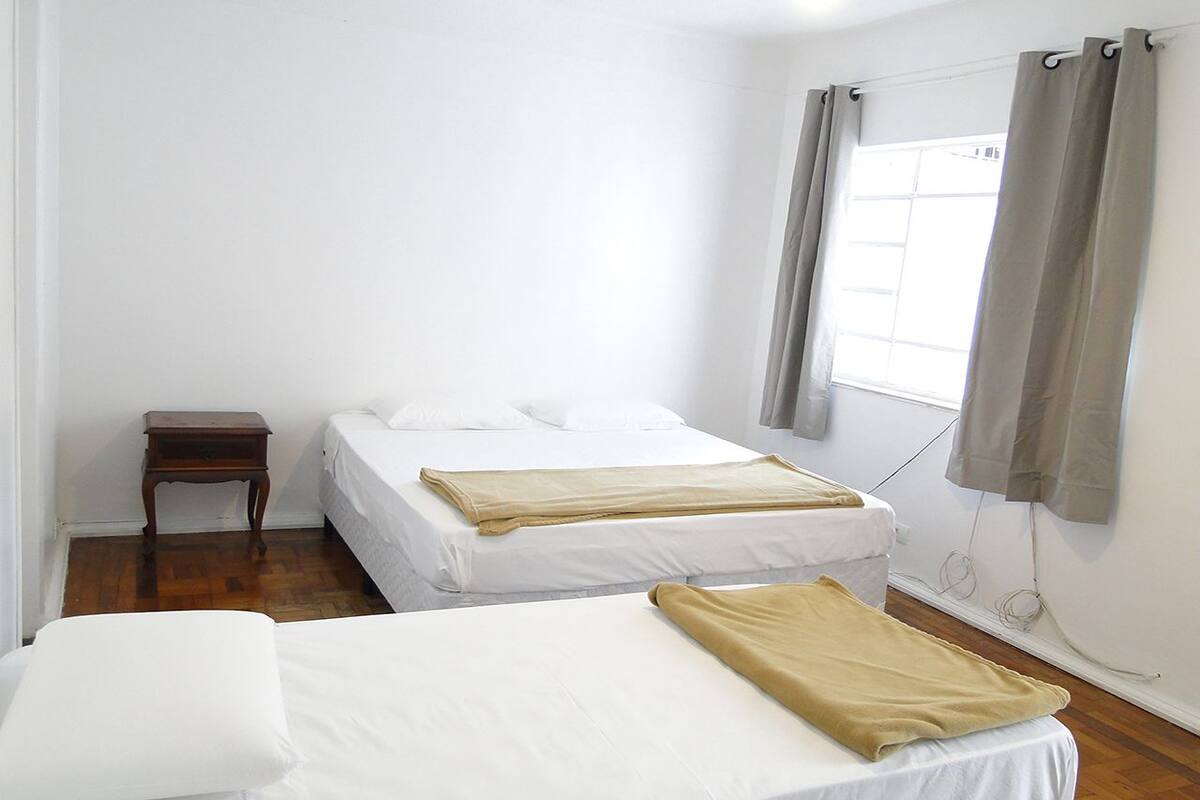 Ein Zimmer mit zwei Betten, einem kleinen Nachttisch aus Holz und einem Fenster mit Vorhängen.